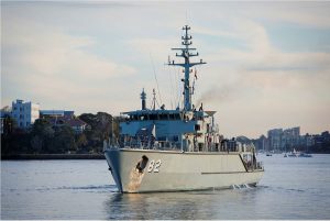 A gray naval ship, marked with the number 82, sails in calm waters near a coastal city. Residential buildings and trees are visible in the background under a partly cloudy sky.