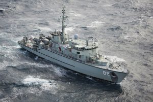 A military patrol vessel with the number 82 on its side navigates through choppy ocean waters. The ship has a large radar on top and multiple antennas, suggesting a focus on surveillance and communication. The sky is overcast.
