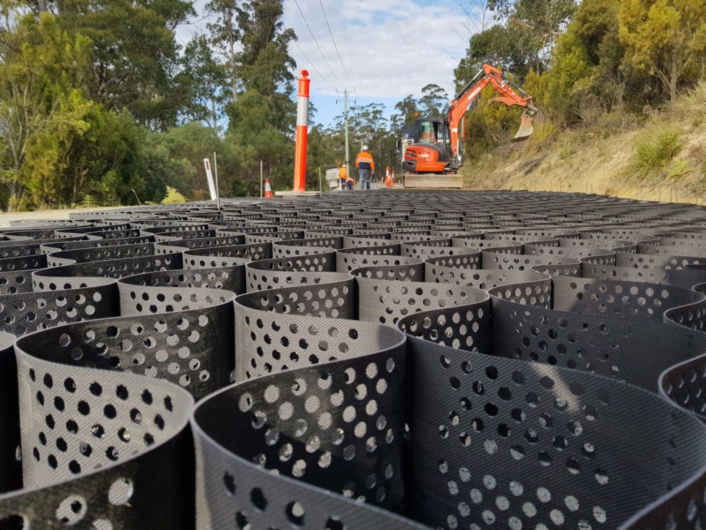 A construction site with a close-up of black geocells placed on the ground. In the background, workers and an orange excavator are near a dirt hill, surrounded by trees and utility poles under a partly cloudy sky.