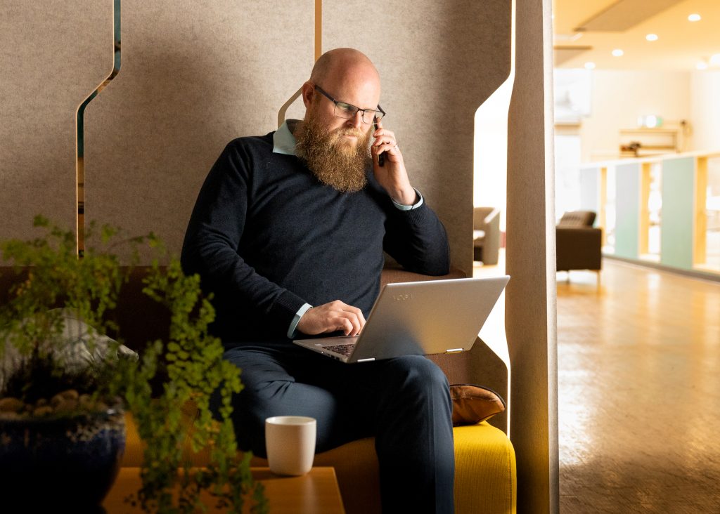 A bearded man sits on a bench in a modern room, talking on a cellphone and using a laptop. A cup and a plant are on the table nearby. The area is softly lit, with a cozy and professional atmosphere.