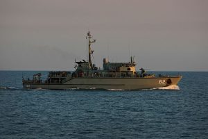 A military patrol boat with the number 82 on its hull navigating through calm sea waters under a hazy sky. The ship is equipped with antennas and various equipment on its deck.