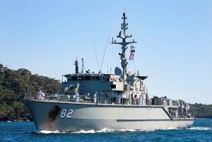 A naval ship marked with the number 82 is sailing on the water. The ship has multiple antennas and flags. Several crew members are visible on deck. The background features a coastline with lush greenery under a clear blue sky.