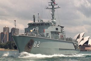 A military ship with the number 82 on its side sails through the ocean near a cityscape. The Sydney Opera House is visible in the background under a partly cloudy sky.