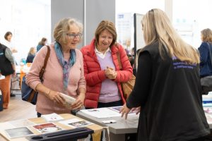 Three women are at a booth at an indoor event. Two women on the left are engaged with the woman on the right, who is presenting materials on a table. People are visible in the background, suggesting a busy atmosphere.