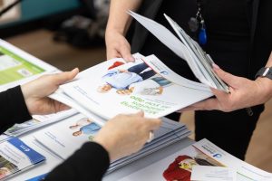 Two people holding and exchanging brochures at a table. The brochures feature images and text on them. One person wears a lanyard, and there are more brochures stacked on the table.