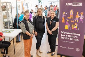 Three people stand smiling beside a banner that says AGE WELL: Networking for seniors in the Huon. The room is filled with people engaged in activities. Tables and brochures are visible in the background.