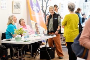 A group of people at a community event with informational tables. One person is smiling and gesturing while others sit at a table covered in pamphlets. A banner in the background reads NDIS. The atmosphere is friendly and engaging.