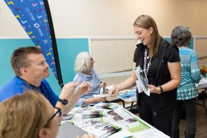 People are gathered at a table in a community event setting. A woman is handing a booklet to a man, while others are seated, engaging in conversation. Pamphlets and brochures are spread on the table, and a banner is partially visible in the background.