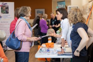 People interacting at a community event with informational pamphlets. A woman in a white top is speaking to another woman holding a pamphlet. The table has brochures, cups, and oranges. Other attendees are visible in the background.