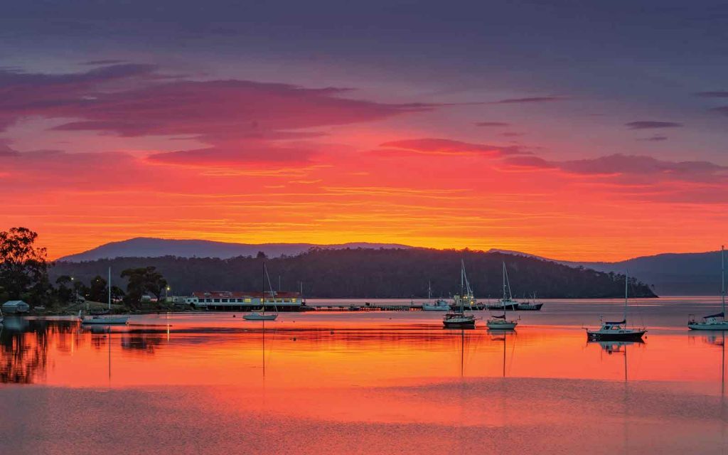 A serene sunset over a calm harbor with several boats anchored. The sky is a vibrant mix of orange, pink, and purple, reflecting beautifully on the water. Hills are visible in the distance, enhancing the tranquil scene.