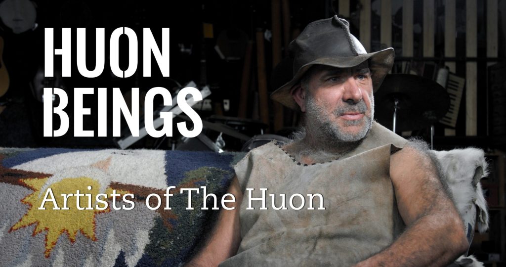 A bearded man wearing a worn leather hat and rustic clothing sits on a patterned couch. Behind him, various musical instruments are visible. Text reads, HUON BEINGS Artists of The Huon.