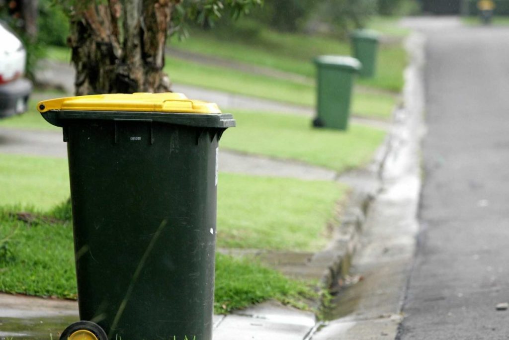 A green wheelie bin with a yellow lid is positioned on a grass verge next to a paved street. In the background, additional bins line the verge at intervals. Trees and parked cars are partially visible further down the street.