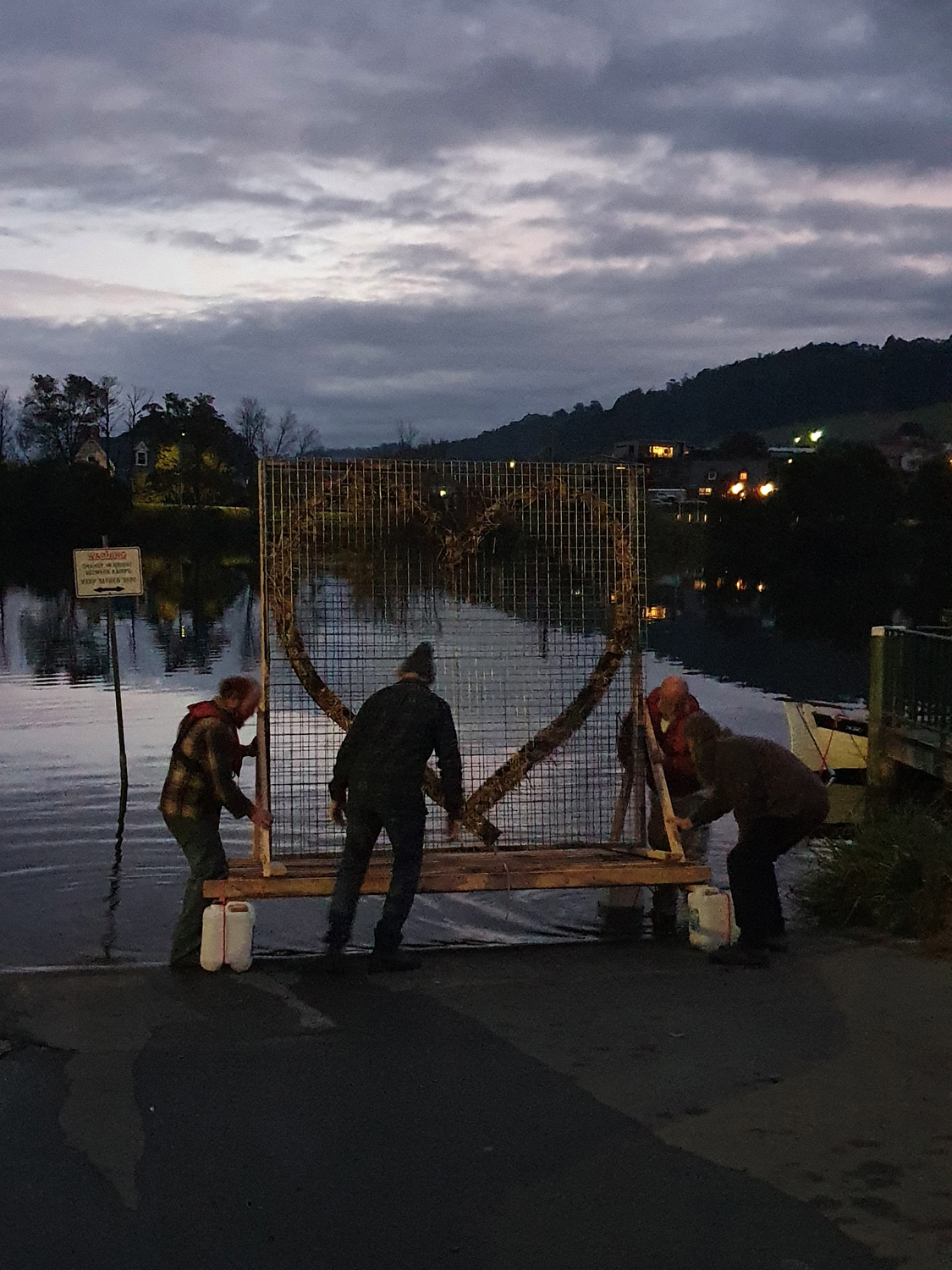 Four people are adjusting a large circular wooden structure on a wire frame by the edge of a lake at dusk. The sky is cloudy, and there are hills and buildings in the background.