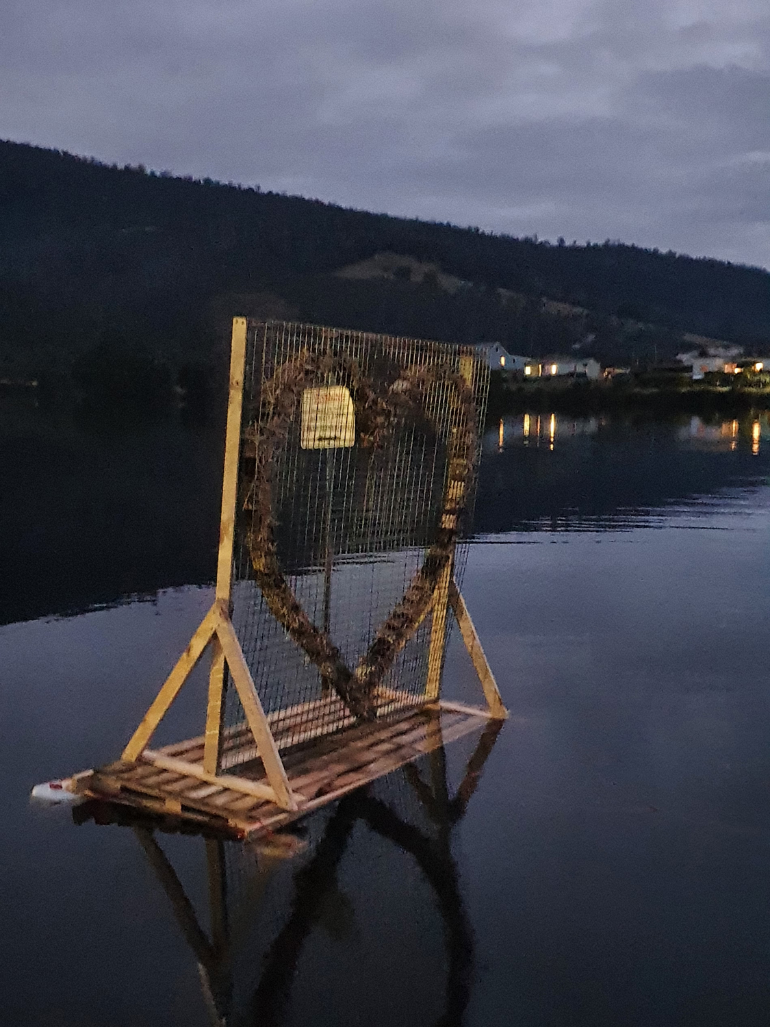 A wooden and wire structure shaped like a heart stands on a pallet floating on a calm body of water. Hills and lights from buildings are visible in the background under a cloudy sky.