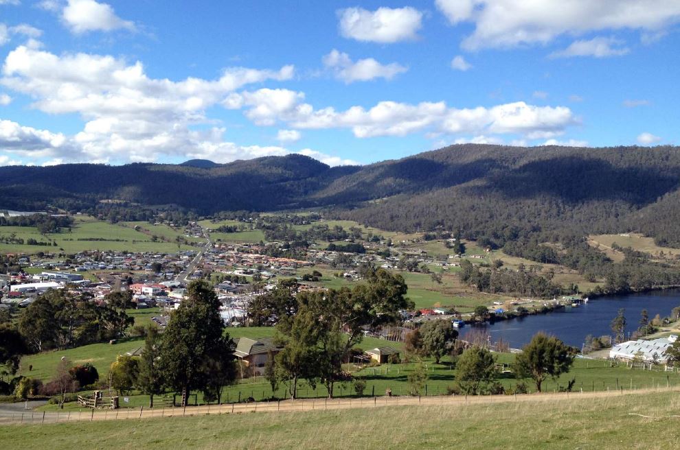 A scenic view of a rural valley with small town buildings, surrounded by lush green fields and forests under a blue sky with scattered clouds. A river flows along the right side, nestled between rolling hills and mountains.