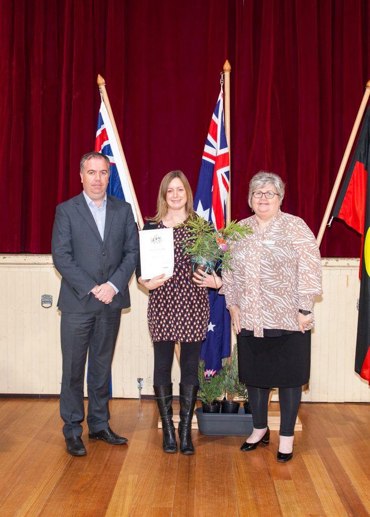 Three people stand on a stage. The person in the middle holds a certificate and a plant. Three flags are displayed behind them. The backdrop is a dark red curtain, and the floor is wooden.