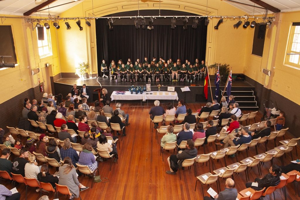A hall filled with seated people facing a stage where a group of children in green uniforms are singing. Decorations include national flags and a table with awards or certificates. The hall has wooden floors and a high ceiling.