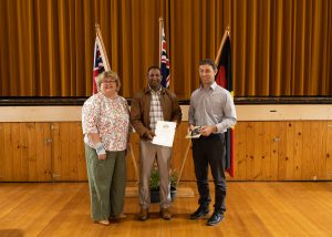 Three people stand in a hall with wooden flooring and flags in the background. The person in the center holds a certificate. The two others stand on either side, smiling.