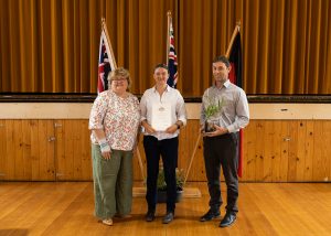 Three people stand indoors on a wooden floor. The person in the middle holds a certificate. Flags stand behind them against a brown curtain. The person on the right holds a small plant.