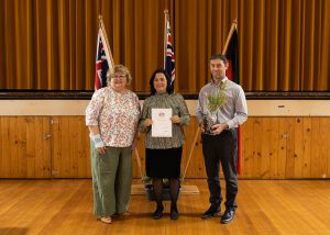 Three people stand on a wooden stage, smiling. The person in the center holds a certificate, and the person on the right holds a small potted plant. Behind them are flags and a brown curtain.