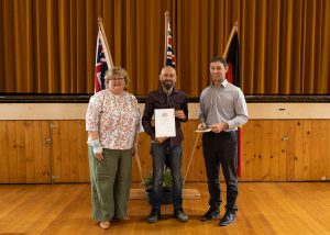 Three people stand on a wooden floor in front of flags. The person in the middle holds a certificate. They are all smiling and dressed in casual and semi-formal clothing. The background features wooden paneling and a curtain.