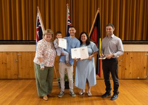 A group of five people, including a child, stand indoors holding certificates. Flags are in the background, and a gold curtain hangs behind them. They are all smiling and standing on a wooden floor.