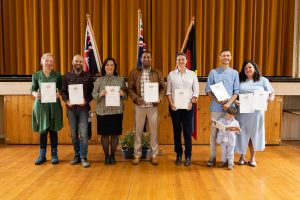 A diverse group of seven people stands on a stage holding certificates. Behind them are three flags. One person is holding a child. The setting suggests a formal or celebratory event.