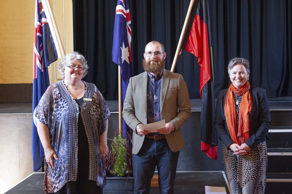 Three people stand on a stage in front of Australian and Aboriginal flags. The individuals, two women and one man, are dressed in formal attire. The man holds a piece of paper, and the group is smiling at the camera.