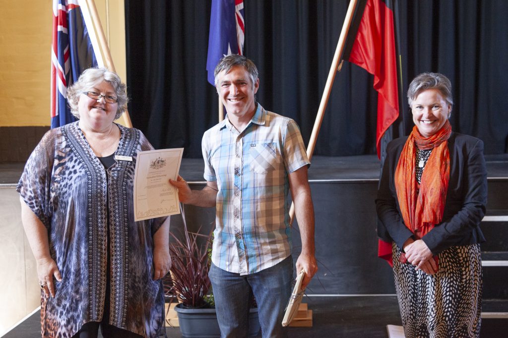 Three people stand on a stage; the woman on the left holds a certificate. Behind them are flags and a potted plant. The group is smiling, and the setting appears formal, possibly a ceremony.