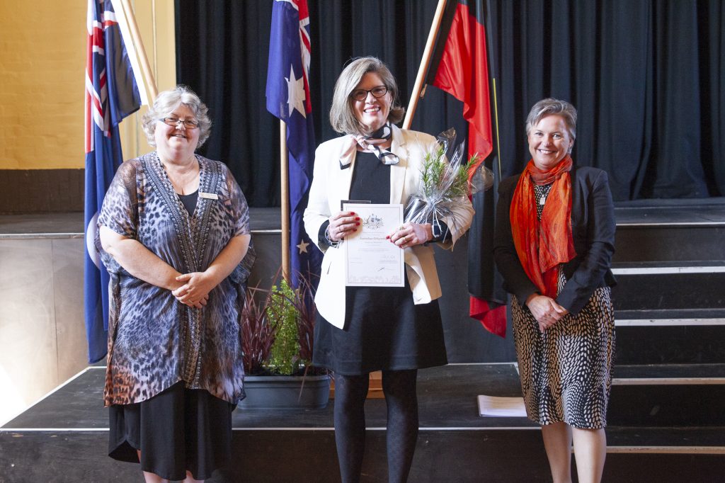 Three women are standing on a stage with flags in the background. The woman in the center holds a bouquet of flowers and a certificate. They are all smiling and wearing formal attire.