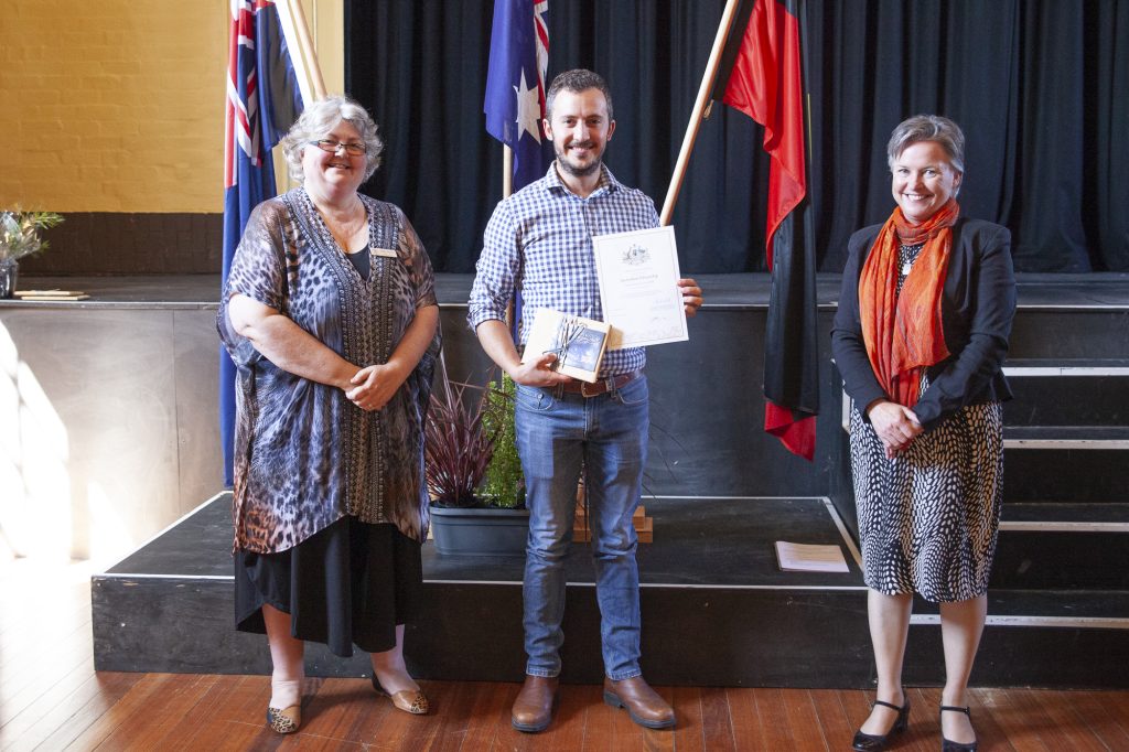 Three people stand on a wooden stage. The man in the middle holds a certificate and a small flag. Two women stand on either side of him. Flags are displayed in the background.