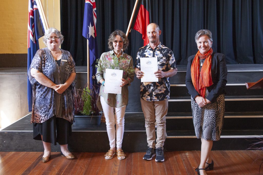 Four people stand on a wooden floor in front of a stage with Australian flags. Two individuals hold certificates. The group is smiling and dressed in semi-formal attire.