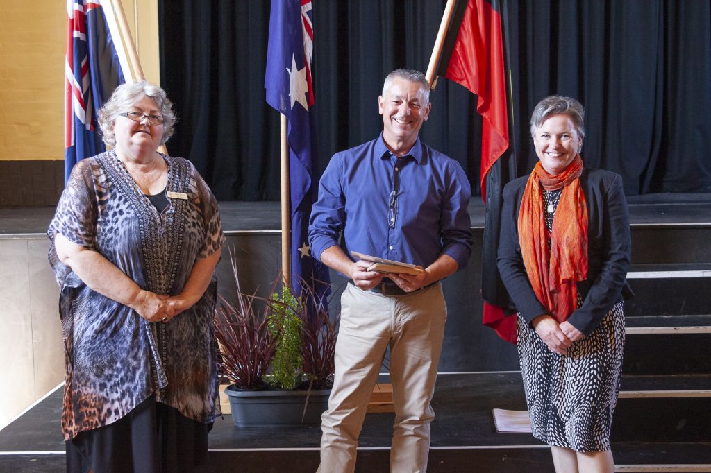 Three people stand indoors in front of several flags, smiling at the camera. The man in the center holds a plaque. The woman on the right is wearing a red scarf, and the woman on the left is wearing a patterned dress.