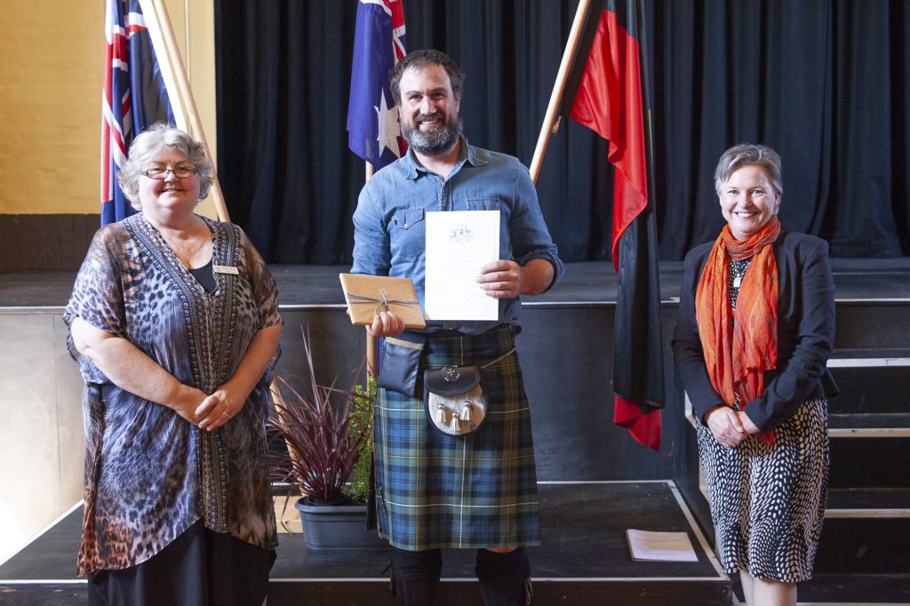 Three people stand on a stage with flags in the background. The person in the center, wearing a kilt, holds a certificate and small box. The two others, a person on the left in a patterned top and a person on the right in a scarf, smile toward the camera.