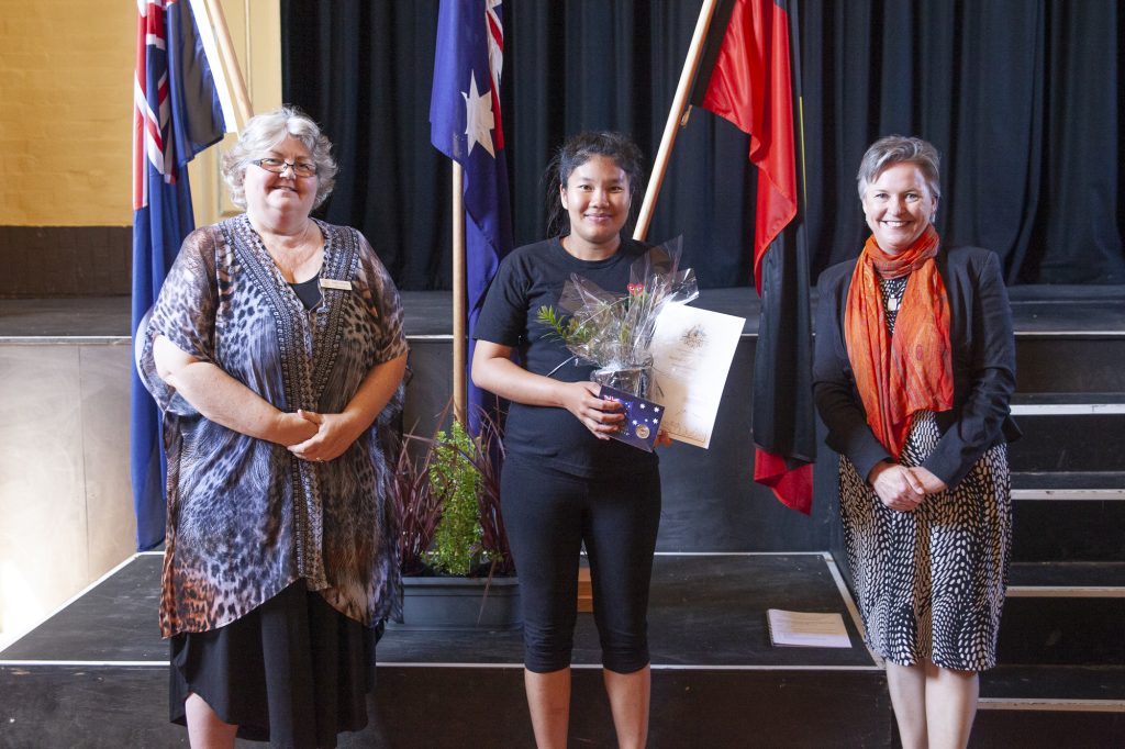 Three women stand on a stage, with two of them holding bouquets and a certificate amidst several national flags. The woman in the center holds flowers and a certificate. They are dressed in formal attire.