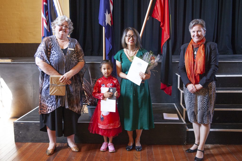 Four people stand on a stage in front of three flags. A woman in a patterned dress, a child in a red dress, another woman in a green dress holding certificates, and a woman in a black and white skirt with an orange scarf smile at the camera.