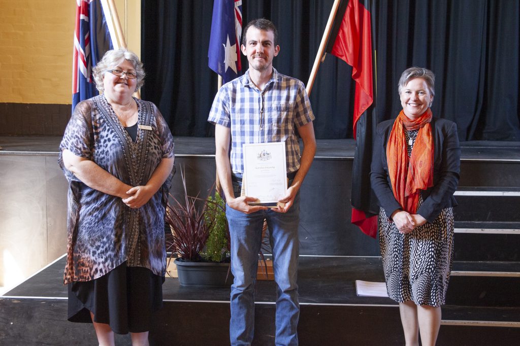 Three people stand on a stage with flags behind them. The person in the middle holds a certificate. The two individuals on either side are smiling, wearing formal attire.
