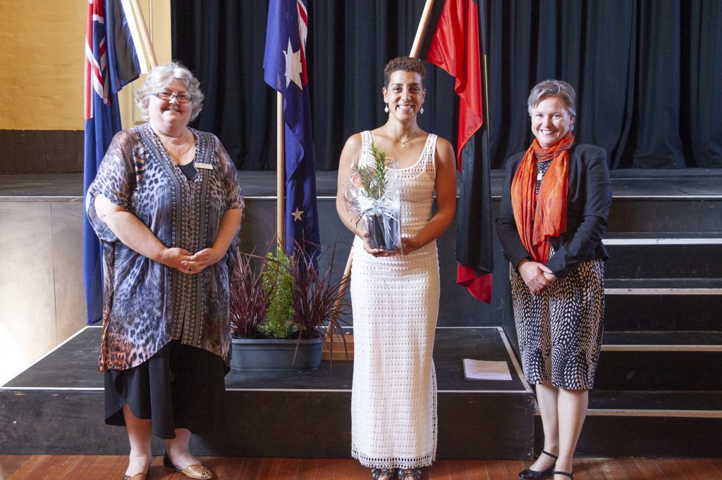 Three women stand indoors, smiling. The woman in the center holds a small potted plant. Behind them are flags, including the Australian flag. They are on a stage with a black curtain backdrop.