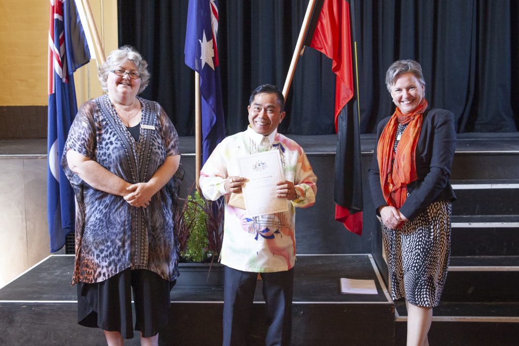 Three people stand on a stage with a man in the center holding a certificate. Two women are beside him, one on each side. They are smiling. Flags are displayed in the background.