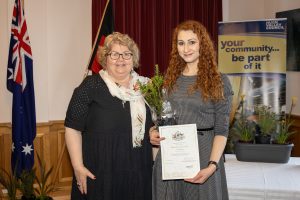 Two women standing indoors; one holds a certificate and a small plant. An Australian flag and a banner saying your community... be part of it are in the background. They are smiling and formally dressed.