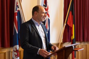 A man in a suit stands at a wooden podium, speaking. Behind him are red curtains and several flags, including a Union Jack and a flag with black, red, and yellow stripes.