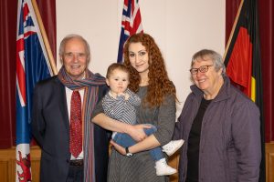 A group of four people, including a man in a suit and scarf, a woman holding a young child, and another woman wearing glasses, stand in front of three flags draped in the background indoors.
