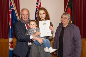 A group of four people pose indoors. A man in a suit holds a toddler while a woman holds a certificate; another woman stands beside them. Two flags are visible in the background.