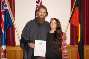 A man and woman stand smiling in front of Australian flags. The man holds a certificate, and both are dressed casually. The background includes red curtains and a wooden panel.