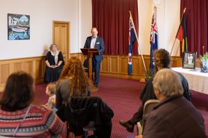 A man speaks at a podium in a room with red curtains. Three flags are on display behind him. Attendees sit facing him, including a woman with a child. A table with framed pictures and items is to the right.