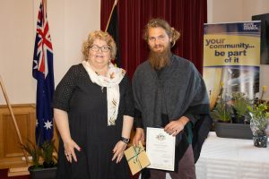 Two people stand indoors near an Australian flag and a banner. One is holding a certificate and gift package. They are both smiling. A table with potted plants is in the background.