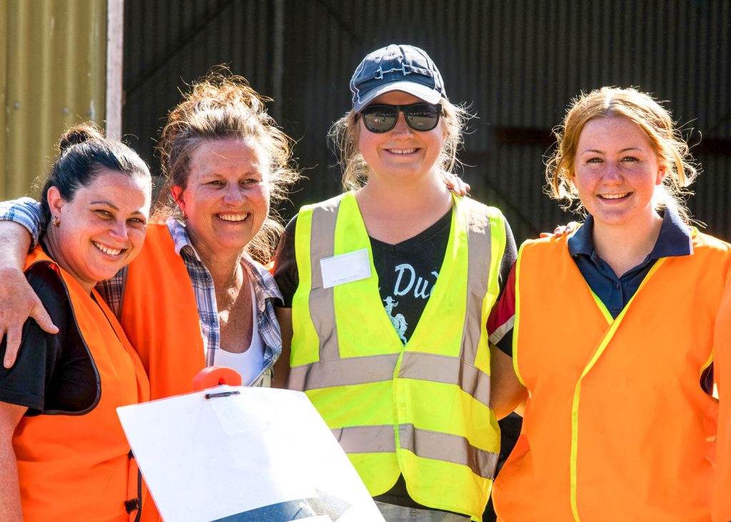 Four people stand closely together, smiling at the camera. They are wearing brightly colored safety vests, and one person is holding a whiteboard. They appear to be outdoors in a sunny environment.