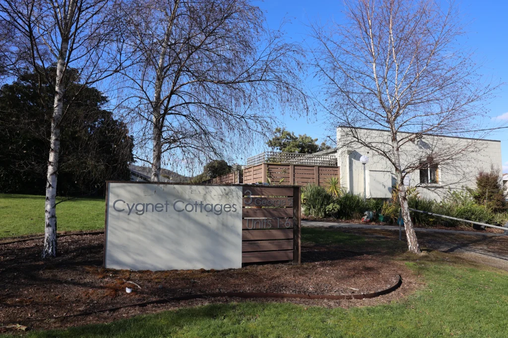 A sign for Cygnet Cottages stands on a grassy area with bare trees nearby; behind it is a brown wooden fence and a light-colored building under a clear blue sky.