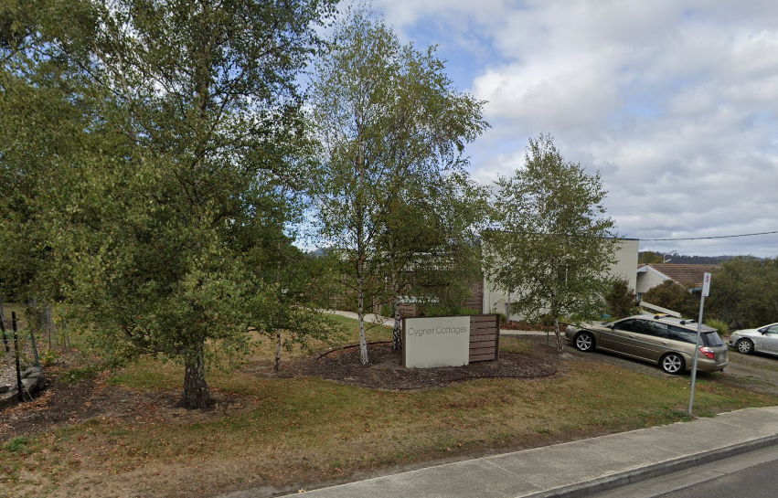 A sign reading Crystall Cottage stands among trees and landscaping near a parking lot with several cars, beside a sidewalk and road under a partly cloudy sky.