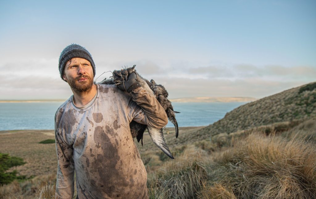 A rugged man in a beanie and dirty clothes stands on a grassy hill overlooking a body of water. He carries a bundle of dead birds over his shoulder. The sky is cloudy, and rolling hills stretch into the distance.